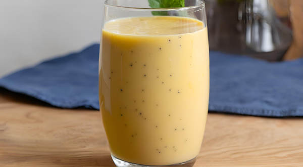 Fotografia de um copo de vidro grande com vitamina de maracujá. No fundo tem um pano azul escuro, que está sobre uma mesa de madeira.