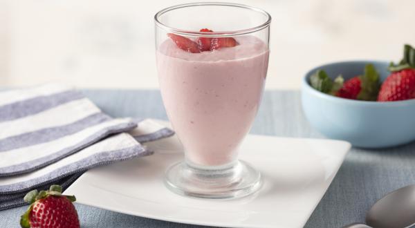 Fotografia em tons de azul e rosa de uma bancada com um prato quadrado, sobre ele um copo com o creme de morango e pedacinhos de morango. Ao redor um paninho listrado azul e branco e um potinho com morangos.